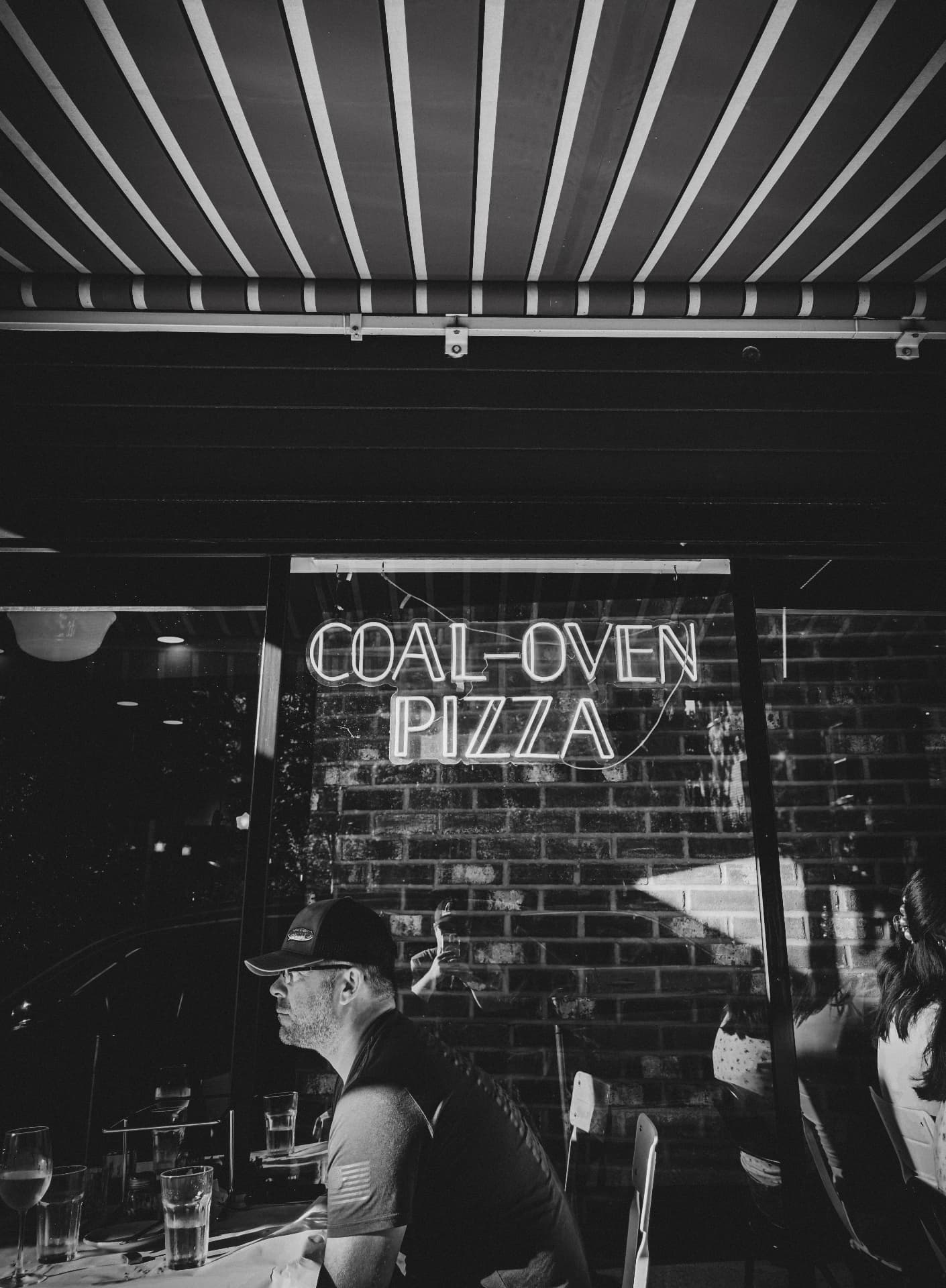 Man seated outside a coal-oven pizzeria at night beneath a striped awning and neon sign, black and white