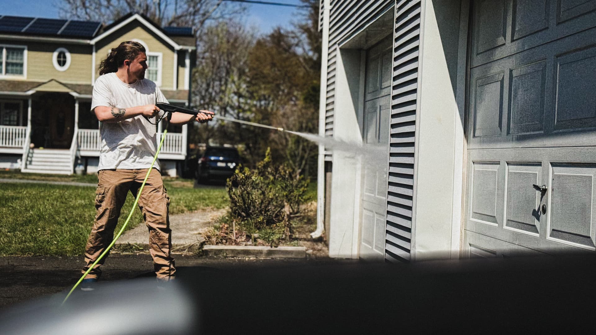 Outdoor cleaner pressure-washing a garage beside a house, with a bright green hose