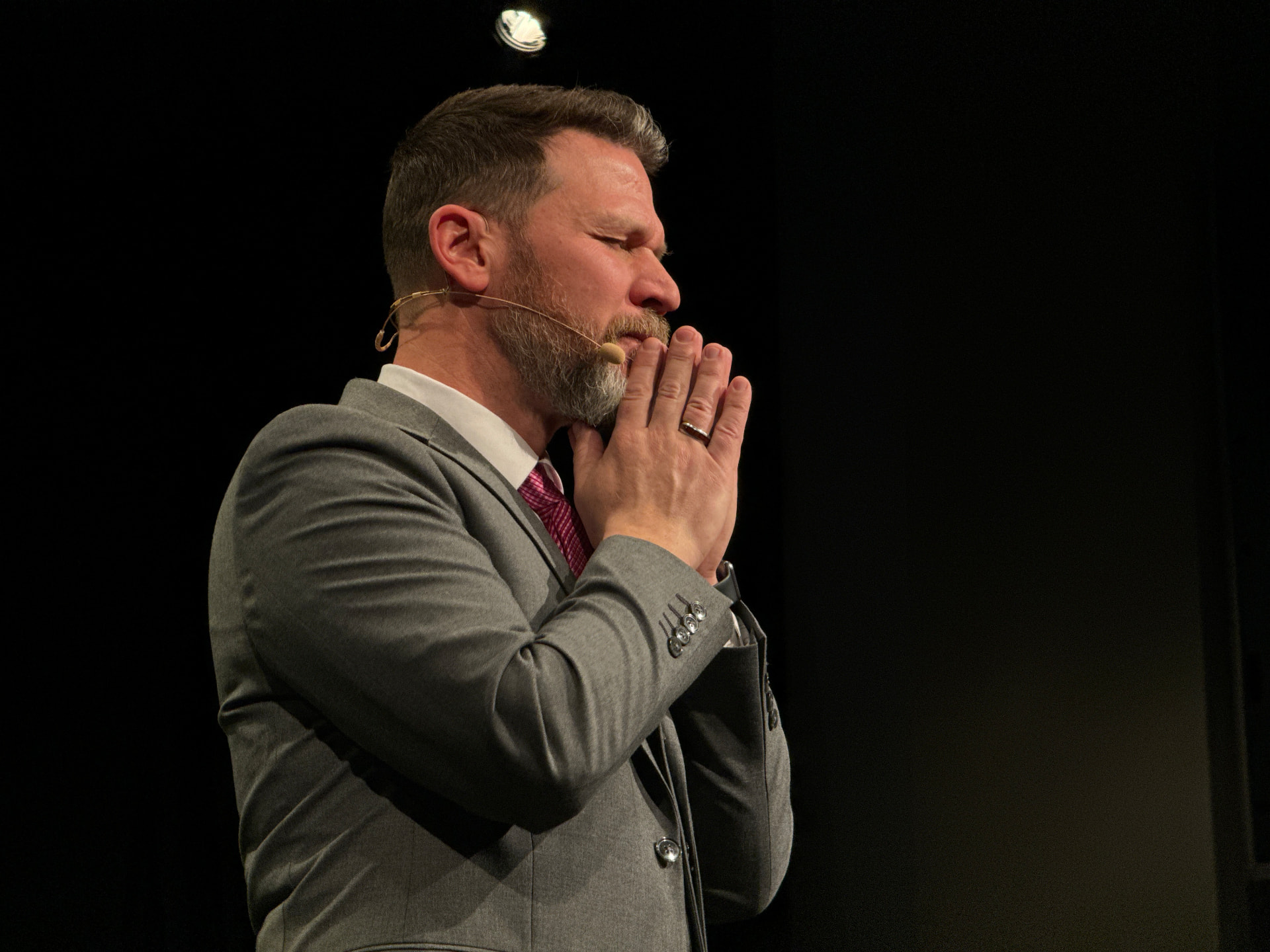 Man in a gray suit with clasped hands and a headset microphone on a dark stage
