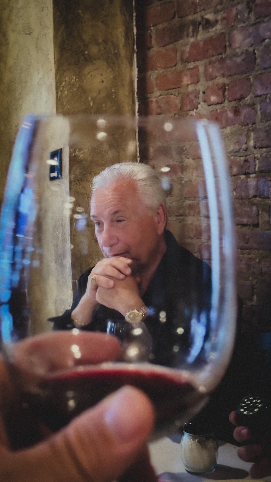 Older man seated at a table, framed through a red-wine glass in the foreground