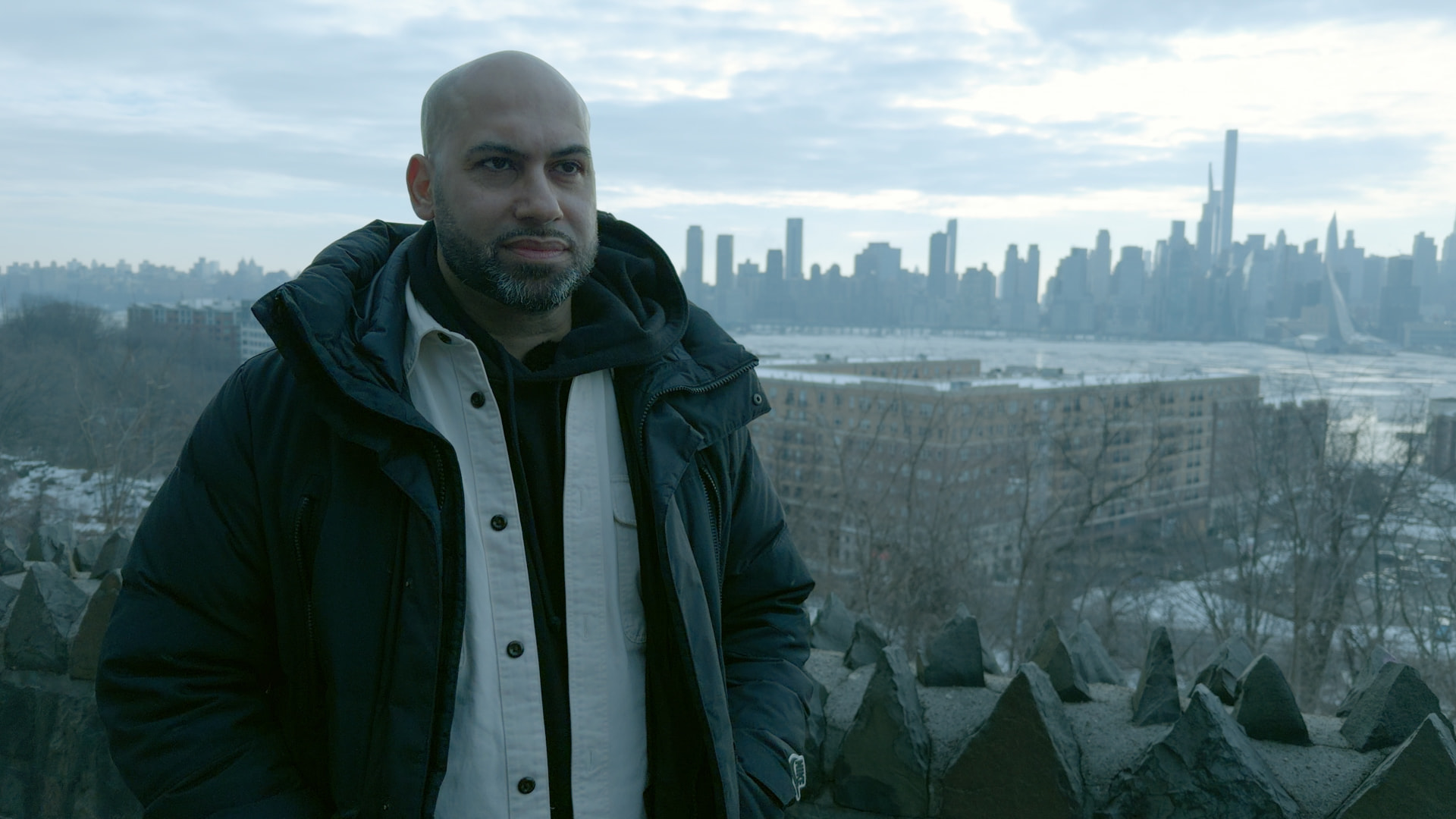 Portrait of a man standing on rocks beside a river with the New York City skyline behind him