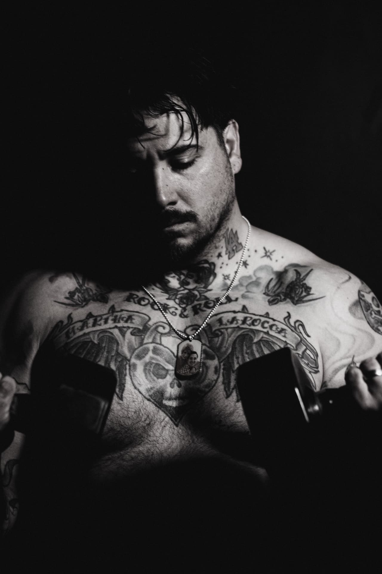 Black and white portrait of a tattooed lifter holding dumbbells with a photograph pendant around his neck